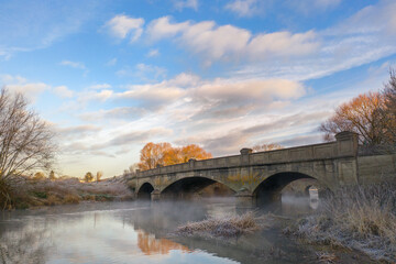 Fototapeta premium bridge crossing a calm, misty river on a cold winter morning, with frost-covered grasses and leafless trees under a pastel sky filled with soft clouds and gentle golden light