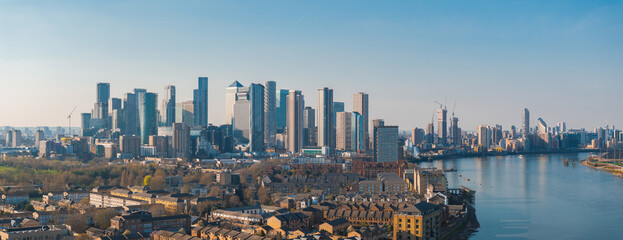 Naklejka premium Aerial perspective of London's Canary Wharf skyline with modern skyscrapers, the River Thames, and surrounding urban neighborhoods under clear skies.