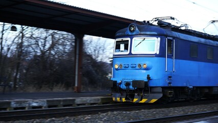 Blue electric locomotive passing through a rural train station with blurred motion, highlighting...