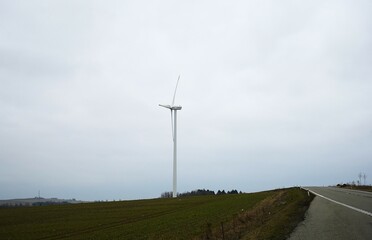 A tall wind turbine stands against a cloudy sky beside a gently curving road through farmland.