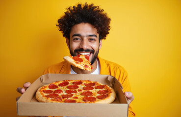 Happy young Indian man eating delicious pizza. Curly guy student holds open box, biting piece of fast food, smiling. Male enjoys meal, pizza slice isolated on yellow background. Junk food