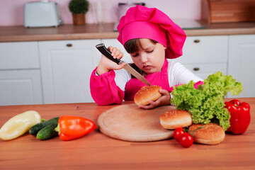 Funny little beautiful child girl in a chef costume prepares burgers while sitting at a table in the kitchen. Healthy eating. Child is cutting sandwich buns with a knife.	