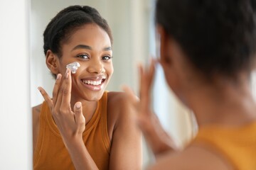 Young woman applying moisturizing cream on face while smiling in mirror, daily skincare routine for healthy glowing skin, natural beauty self-care lifestyle indoors