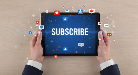 Holding Tablet with Subscribe Message Surrounded by Social Media Icons on Wooden Table