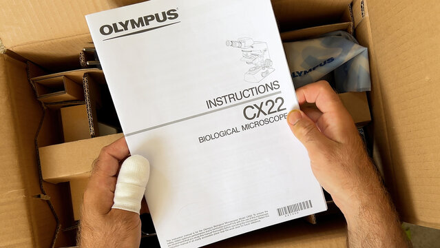 Paris, France - Apr 9, 2024: A person with a bandaged thumb is unboxing a cardboard package containing an Olympus CX22 biological microscope, holding its instruction manual above the packing materials
