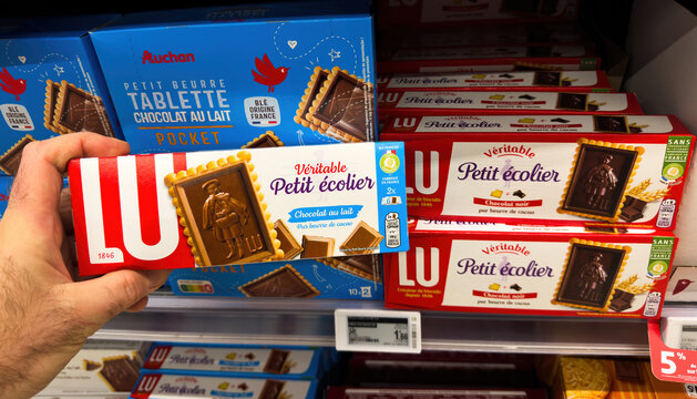 Paris, France - Feb 13, 2025: A hand holds a pack of LU Veritable Petit E colier chocolate biscuits with updated 2025 packaging in front of store shelves including milk and dark variants