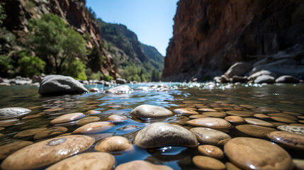 A close-up of the Merced River with smooth stones in the water, framed by towering cliffs and a clear blue sky.