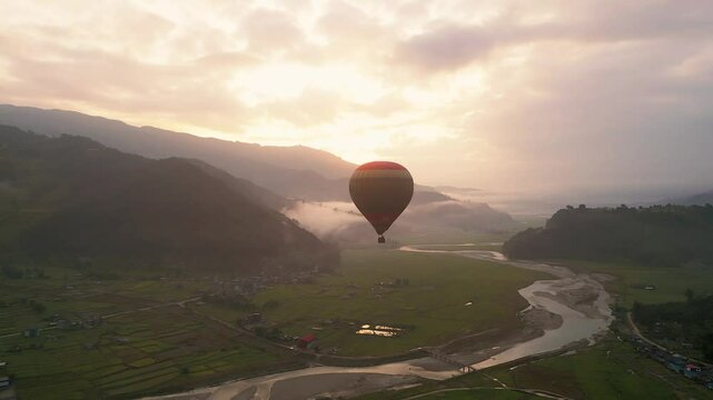 Hot air balloon flying over misty valley at sunset, with green fields, river, and mountains.