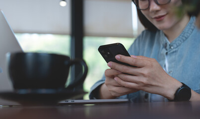Young woman sitting in coffee shop, using mobile phone with laptop computer on table. Asian freelancer online working  on laptop and using smartphone, surfing the internet at cafe, felance lifestyle