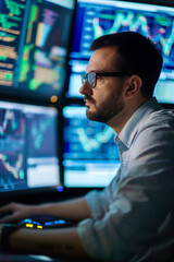 Fototapeta premium A focused man with glasses analyzes data on multiple monitors filled with graphs. He's in a dark room, typing on a keyboard. He seems to be working or researching technical graphs.