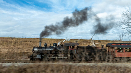 Obraz premium A vintage steam locomotive puffs black smoke as it moves along the tracks in a scenic countryside setting. The overcast sky adds a moody atmosphere to the scene.