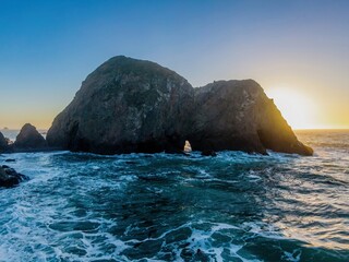 Coastal rock formation with a natural archway is silhouetted by the setting sun, as waves crash...