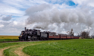 A vintage steam locomotive rolls along a grassy track, billowing smoke against a picturesque rural backdrop of fields and overcast skies. The scene captures the essence of classic train travel.