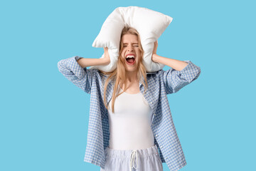 Stressed young woman with pillow in pajamas suffering from insomnia on blue background