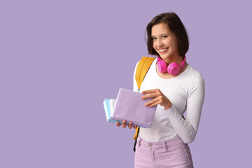 Beautiful female student with headphones, backpack and books on lilac background