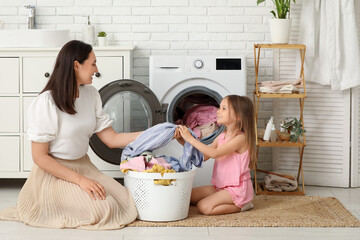 Young woman and her daughter pulling out fresh clothes from washing machine in laundry room