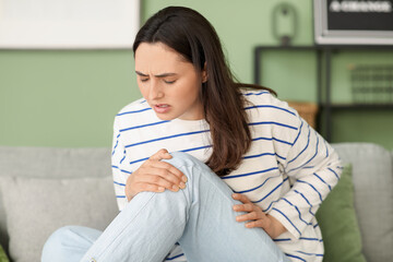 Young woman suffering from pain in knee on sofa at home
