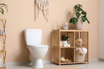 Interior of restroom with ceramic toilet bowl, houseplants and shelving unit near beige wall