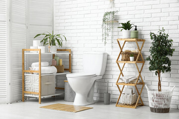 Interior of modern restroom with ceramic toilet bowl, houseplant and shelving unit near white brick...