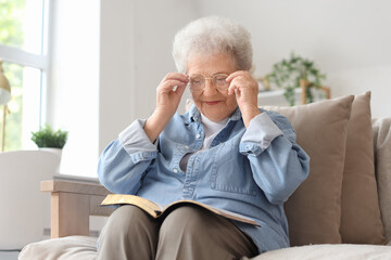 Senior woman with Bible putting on glasses at home