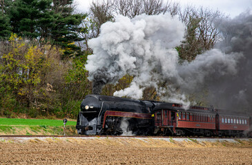 A vintage steam locomotive travels along countryside tracks, releasing clouds of black smoke into the crisp autumn air. Colorful trees provide a backdrop as the train moves forward, evoking nostalgia.