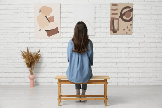 Young woman looking at paintings on white brick wall in art gallery, back view