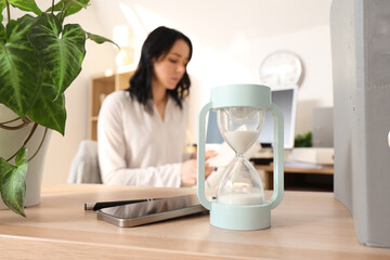 Hourglass with mobile phone on table against businesswoman working in office, closeup