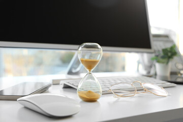 Hourglass with eyeglasses and computer on desk in office, closeup