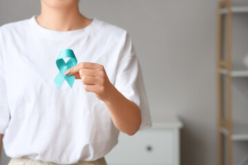 Young woman holding turquoise ribbon at home, closeup. Cervical Health Awareness Month