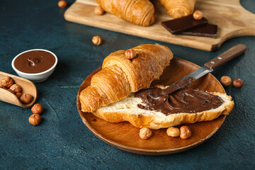 Wooden plate of sweet croissant with chocolate spread and hazelnuts on dark background