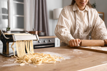 Young woman with rolling pin, pasta machine and dough near table in kitchen