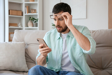 Shocked young man with eyeglasses using mobile phone on sofa at home