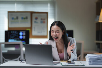 Fototapeta premium Asian businesswoman is expressing excitement and joy, celebrating a successful outcome or achievement while working on her laptop in a modern office environment