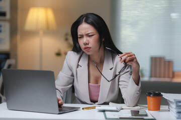 Young professional woman taking off glasses and looking at laptop with concerned expression, working late in office with calculator, paperwork, and coffee on desk