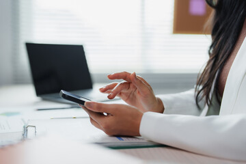 Fototapeta premium Businesswoman scrolling through her smartphone while working at a desk in a bright, modern office, surrounded by a laptop and important documents, focusing on productivity and communication