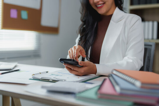 Businesswoman smiling and using smartphone while working on financial reports and business documents in modern office, managing her tasks and staying connected - Powered by Adobe