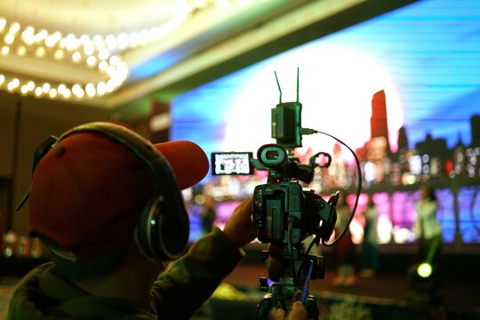 Cameraman filming event with large screen in background.  Person wearing headphones operating professional video camera.