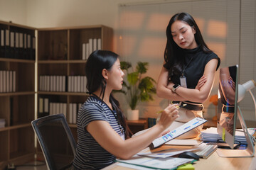 Two young asian businesswomen are discussing work in an office at night, with one pointing at a chart and the other listening with arms crossed