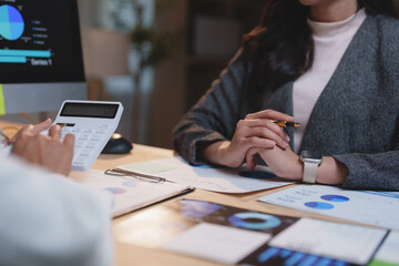 Two businesswomen are collaborating on a project, analyzing financial charts, using a calculator, and discussing data in a modern office setting