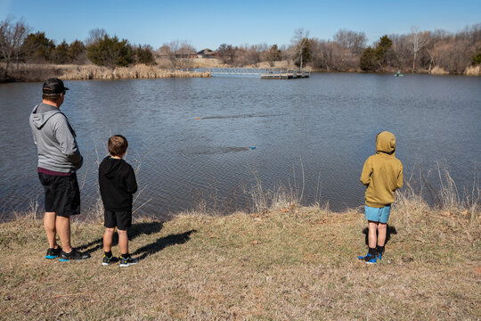 Three people watch remote control boats on a calm lakeside shore