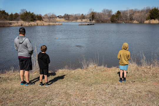 father with two sons driving remote control boats on a lake