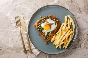 Tasty hot Schnitzel Holstein served with a fried egg, anchovy and lemon parsley caper sauce on top close up on the plate on the table. Horizontal top view from above