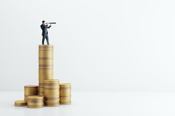 Businessman standing on stacked gold coins looking through telescope on light background,...