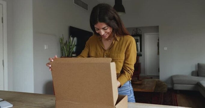 Woman stand at table opening large cardboard box with positive expression, enjoy received gift, got surprise or bought goods for home. Electronic commerce, webstore client, express transport services