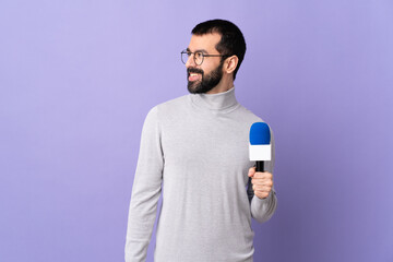 Adult reporter man with beard holding a microphone over isolated purple background looking to the...