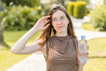 French girl with glasses holding a bottle of water at outdoors having doubts and with confuse face...