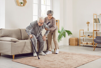 Senior Caucasian woman helping her elderly gray-haired man husband to walk with walking cane standing in the living room at home. Retired couple take care of each other in retirement.