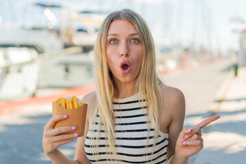 Young blonde woman holding fried chips at outdoors surprised and pointing side
