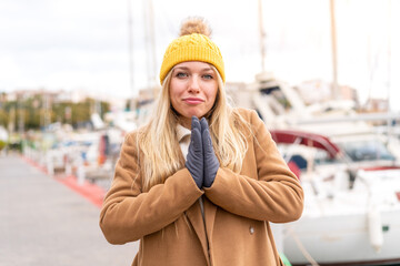 Young pretty blonde woman wearing winter jacket at outdoors keeps palm together. Person asks for...