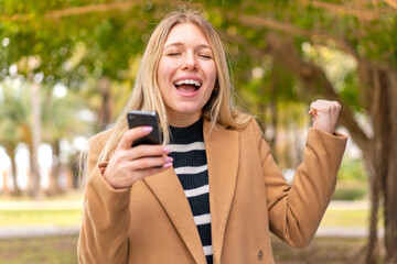 Young pretty blonde woman at outdoors using mobile phone and doing victory gesture